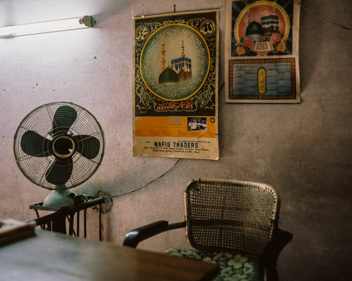 "Office Desk" an inkjet print by Saleem Ahmed. depicts an sparsely furnished office with two colorful posters on the wall. Cityscape, fan, office, urban landscapes. The Print Center