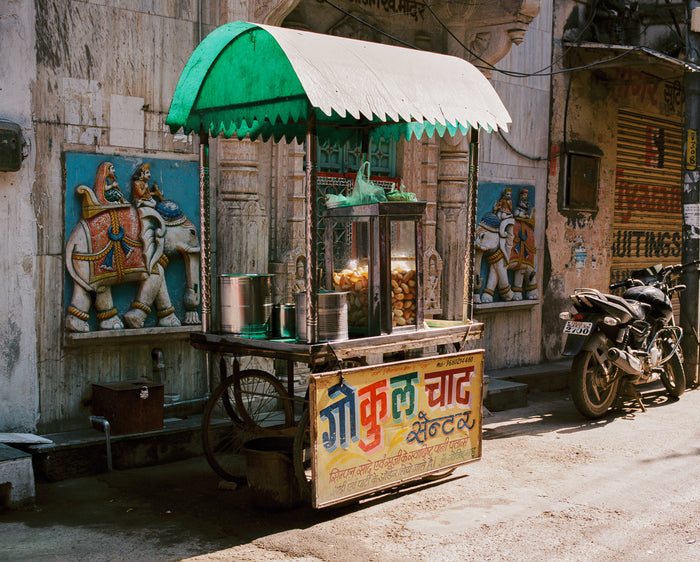 "Pani Puri" an inkjet print by Saleem Ahmed. An organe juice stand sits along astreet infront of murals of elephants and their riders. Color Photography, cityscapes, urban landscapes. The Print Center